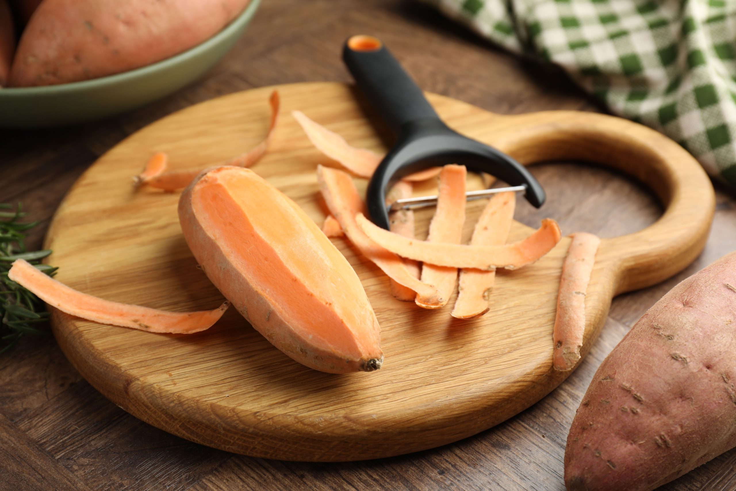 Raw sweet potatoes, peel and peeler on wooden table, closeup